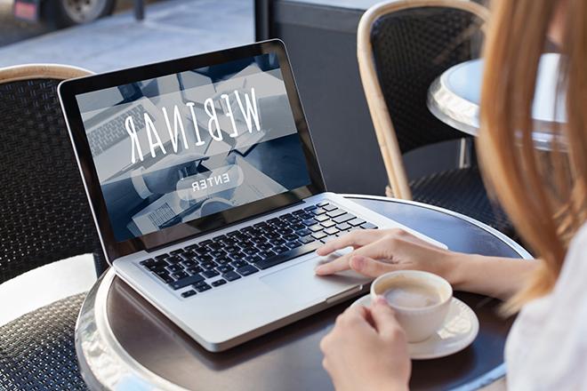 A woman sits at a coffeshop, watching a webinar on her laptop.